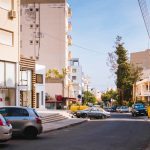 Sunlit street in a Mediterranean city with cars and modern architecture.