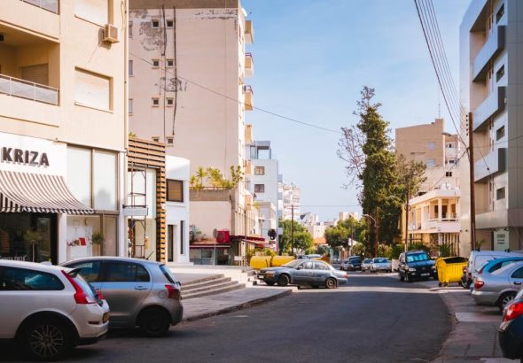 Sunlit street in a Mediterranean city with cars and modern architecture.