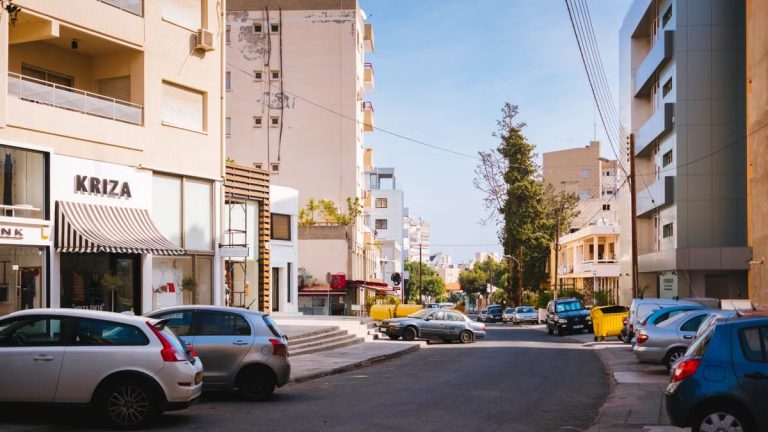 Sunlit street in a Mediterranean city with cars and modern architecture.